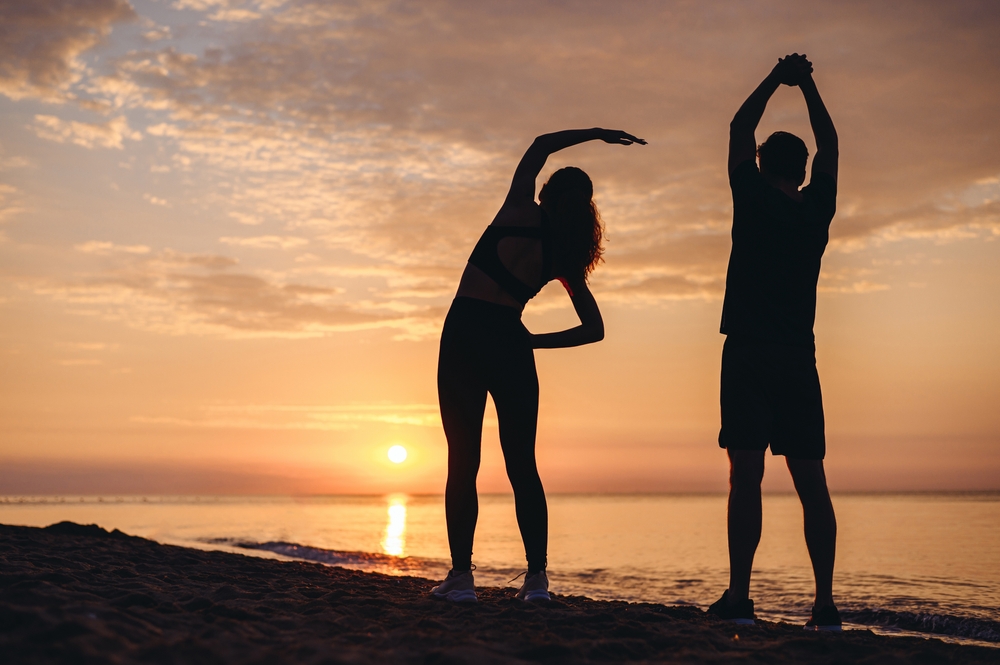 Two people stretching together on the beach at sunrise, symbolizing wellness, flexibility, and recovery through physical therapy – Physical Therapy in Manahawkin, NJ.